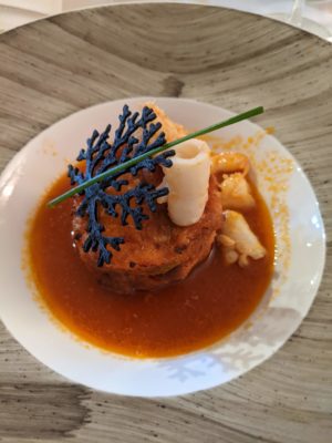 savory bread pudding with seafood served in a fish broth - this is a traditional dish for poor fisherman - the black decoration is some sort of fried cheese thing - there were mussels in the bread pudding and shrimp and a tubular thing in the broth